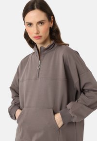 Hellbrauner Zip-Hoodie mit hohem Kragen, elastischen Bündchen und Fronttaschen; hat eine glatte Textur und kontrastierende Nahtdetails.