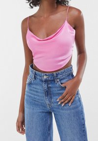 Růžový saténový crop top s volným výstřihem a tenkými ramínky, kombinovaný s džínami s vysokým pasem. Materiál má hladkou, lesklou texturu.