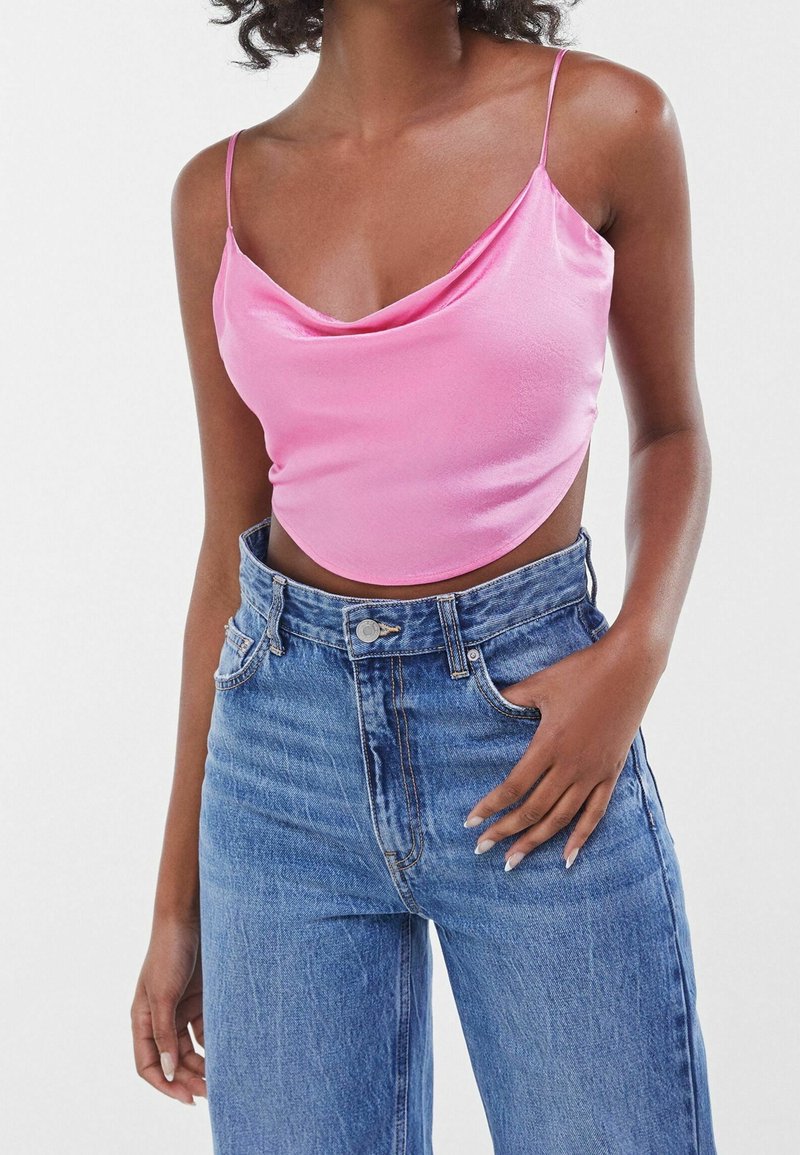 Růžový saténový crop top s volným výstřihem a tenkými ramínky, kombinovaný s džínami s vysokým pasem. Materiál má hladkou, lesklou texturu.
