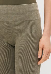 Leggings côtelés vert olive avec une texture douce, dotés d'une large ceinture et de rayures verticales courant sur toute la longueur du tissu.