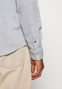 Maglietta a maniche lunghe grigio chiaro con trama, con polsini con bottoni, caratterizzata da due bottoni su ciascun polsino e indossata con pantaloni beige.