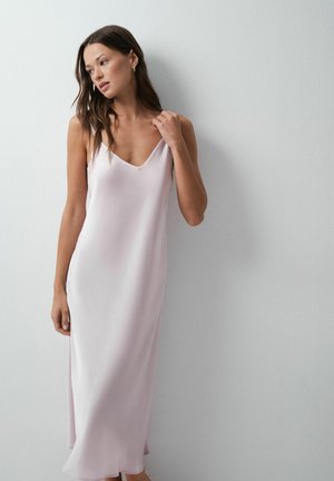 Femme aux longs cheveux bruns portant une robe nuisette légère rose pâle, debout contre un mur blanc uni.