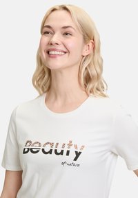 Hvid bomulds t-shirt med teksten "Naturens Skønhed" i en blanding af leopardprint og sort. Rund halsudskæring og korte ærmer.