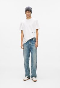 Balta kokvilnas T-krekls ar mazu melnu grafiku, apvienots ar izbalējušiem ziliem platām biksēm un pelēku adītu cepuri. Baltas sporta apavus ar brūniem akcentiem.