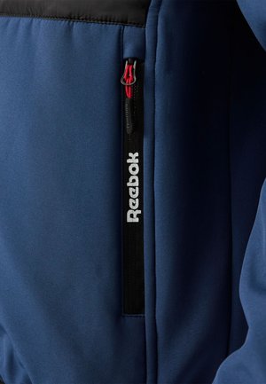 Giacca sportiva blu navy con sezione superiore nera, tasca con cerniera e tirante rosso, e logo "Reebok" bianco lungo la cerniera.