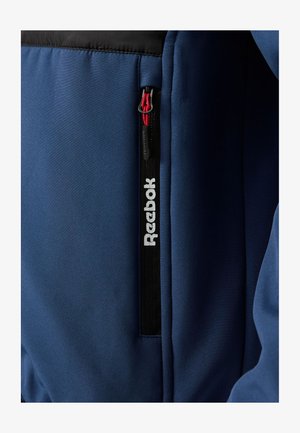 Marineblauw sportjack met een zwart bovenstuk, een zak met rits en een rood trekkoord, en een witte "Reebok"-logo langs de rits.