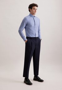 Camicia a righe blu e bianche con colletto alto, abbinata a pantaloni navy su misura e scarpe nere. Tessuto liscio, silhouette aderente.