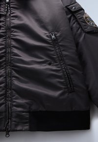 Polstrovaná černá bunda s texturovaným povrchem, s předním zipem, bočními kapsami a žebrovanými manžetami s vyšitým štítkem na rameni.