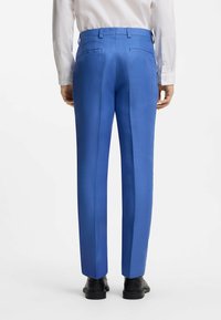 Homme portant un pantalon de costume bleu vif, une chemise blanche et des chaussures noires, vu de dos sur un fond blanc uni.