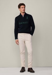 Námořnický svetr na zip s zeleným logem "HACKETT" a číslem "1" v přední části, béžové kalhoty a tmavě hnědé boty. Stojí na jednobarevném pozadí.