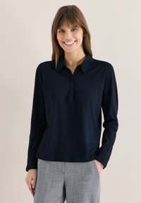 Femme souriante, portant une chemise bleu marine à manches longues avec col et un pantalon à carreaux gris, debout devant un fond clair uni.