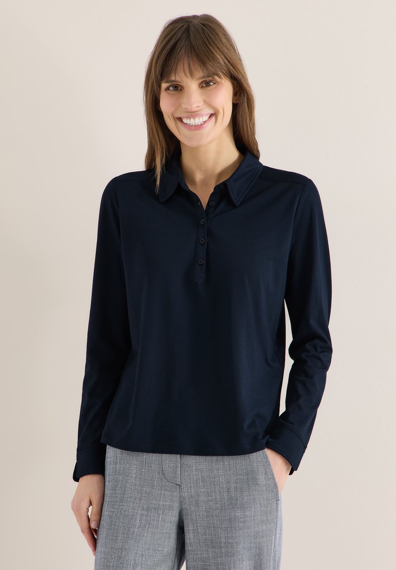 Femme souriante, portant une chemise bleu marine à manches longues avec col et un pantalon à carreaux gris, debout devant un fond clair uni.