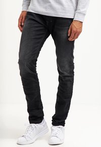 Man wearing black slim-fit jeans, light gray long-sleeve shirt, and white sneakers standing against a white background.