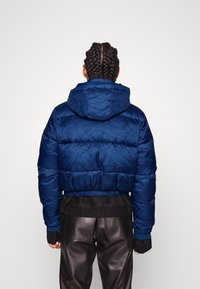 Granatowa, krótka kurtka puchowa z kapturem, z pikowanym wzorem, elastycznym dołem i ściągaczami przy mankietach, nałożona na ciemne skórzane spodnie.