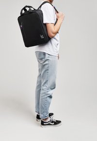 Sac à dos noir de forme rectangulaire, en tissu, avec une poche avant zippée et des accents métalliques colorés. Porté sur une épaule.