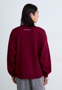 Burgunderfarvet oversized sweater med lange ærmer, ribbede manchetter og en afrundet kant. Har et hvidt "Porsche" logo på ryggen.