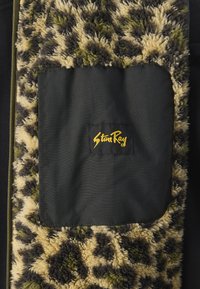 Chlpatá, textúrovaná tkanina so zeleno-čiernym vzorom a čiernou štvorcovou kapsou. Logo "StunRay" v zlatej farbe na čiernom materiáli. Viditeľný detail zipsu.
