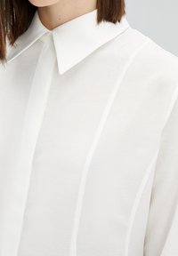 Witte blouse met een gladde, gestructureerde stof. Kenmerkt zich door verticale naden en een klassiek kraagontwerp. Close-upweergave benadrukt de details.