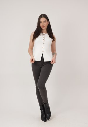 Femme aux longs cheveux foncés portant un haut blanc sans manches boutonné, un jean gris foncé ajusté et des bottines noires, debout devant un fond uni.