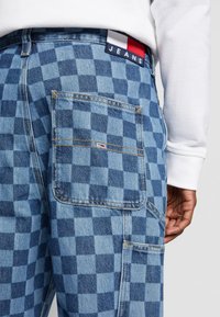 Modré kostkované džíny z denimu mají zadní kapsu, kontrastní švy a značkovou nálepku v červené, bílé a modré barvě poblíž pasu.