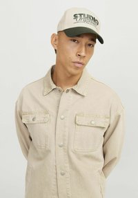 Beige denimjakke med to brystlommer og trykknapper, båret under en hvid og grøn kasket med teksten "STUDIOS Original" trykt på.