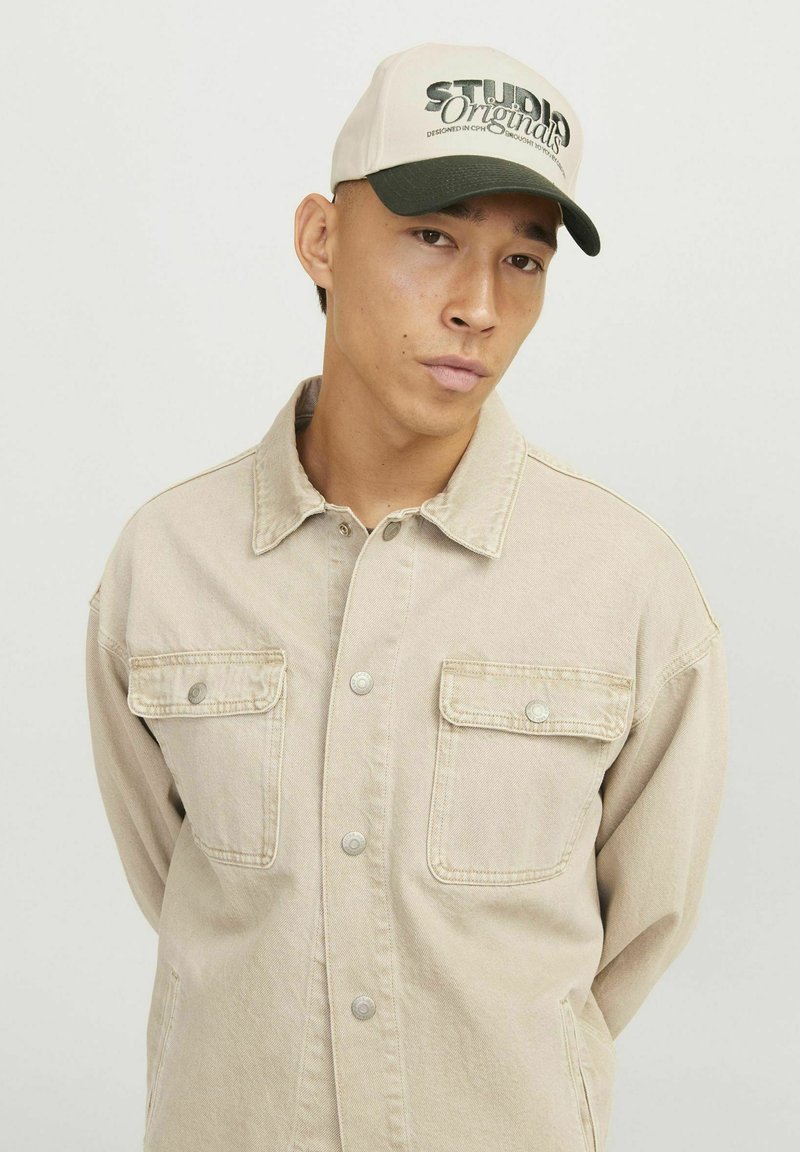 Beige denimjakke med to brystlommer og trykknapper, båret under en hvid og grøn kasket med teksten "STUDIOS Original" trykt på.