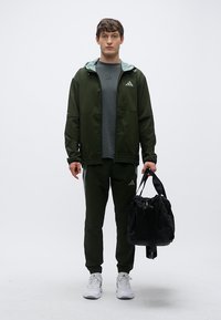 Casaco desportivo verde com capuz, t-shirt cinzenta, calças verdes e ténis brancos; segurando uma mala desportiva preta. Design simples e desportivo.