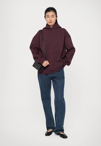 Felpa bordeaux con tasca a marsupio e vestibilità oversize, abbinata a jeans blu a gamba dritta e ballerine nere, completata da una piccola borsa nera.