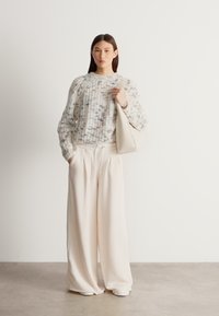 Pulover tricotat alb și gri, cu un model texturat, asortat cu pantaloni bej cu picioare largi și o geantă deschisă la culoare, prezentate pe un fundal neutru.