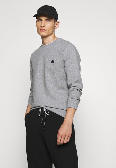 Grijze sweater met ronde hals en zwart merklabel op de borst, gecombineerd met zwarte broek met witte trekkoorden.