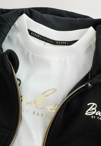 Sudadera con cremallera negra sobre una camiseta de cuello redondo blanca. Texto dorado "Baker by Ted Baker" en ambas prendas, con cuellos acanalados.