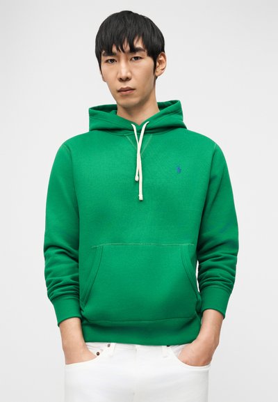 Mladi moški s črnimi lasmi, oblečen v zeleno jopo z belimi vrvmi in belimi hlačami, stoji z rokami v žepih pred enobarvnim ozadjem.