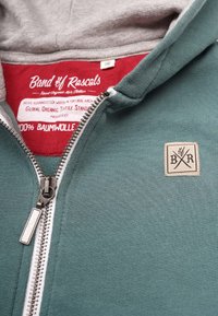 Grön zip-up huvtröja med grå huva, med en vävd etikett, metalldragkedja och mjuk textur. Inuti har den en röd foder med branding.