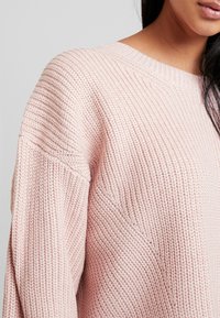 Hellrosa gestrickter Pullover mit einer gerippten Textur, rundem Ausschnitt und abfallenden Schulternähten, der eine lässige Passform und weichen Stoff zeigt.