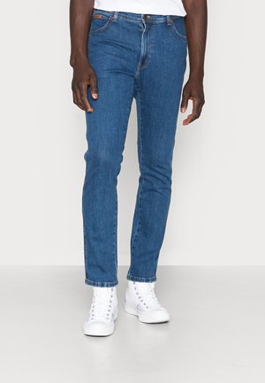 Mann steht und trägt schmal geschnittene blaue Jeans und weiße High-Top-Sneaker vor einem schlichten weißen Hintergrund.