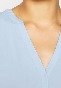 Blouse bleu clair avec un col en V, en tissu lisse, légèrement brillant, agrémentée d'un seul bouton au centre.