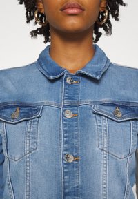 Světle modrá džínová bunda s špičatým límcem, zapínáním na knoflíky a dvěma náprsními kapsami se stříbrnými detaily a oranžovým prošíváním.