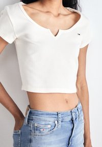 Biała krótka bluzka z prążkowanej tkaniny, z dekoltem w kształcie V i krótkimi rękawami; zestawiona z jasnoniebieskimi szortami dżinsowymi z małą naszywką z logo.