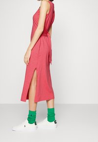 Rosa ärmlös klänning med sidoklipp och knytning i ryggen, kombinerad med vita sneakers och gröna kraftiga strumpor. Slät textur, avslappnad passform.