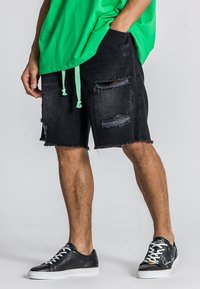 Pantalones cortos de mezclilla negros con detalles rasgados y dobladillo deshilachado, combinados con zapatillas negras que tienen cordones de colores y suelas de goma blanca.
