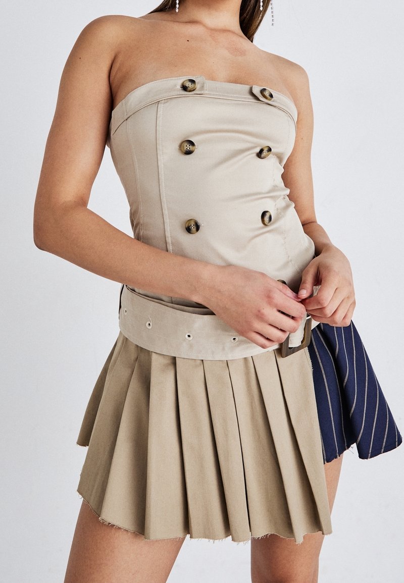 Mujer con top beige sin tirantes con botones, cinturón ancho y falda plisada, sosteniendo una tela a rayas en azul marino y beige.