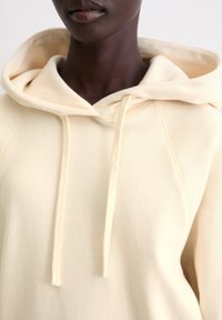 Crème-kleurige hoodie in zachte stof, met een koord, een gevoerde capuchon en subtiele stiksels. Relaxte pasvorm, minimalistisch ontwerp.