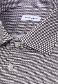 Chemise habillée à col avec un petit motif géométrique marron et blanc, deux boutons blancs visibles et une étiquette "seidensticker" à l'intérieur du col.