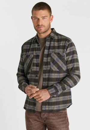 Man with short hair wearing a green and gray plaid button-up shirt over a brown shirt, posing with hands clasped in front.