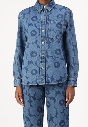 Chemise et pantalon en denim avec grands motifs floraux bleu foncé, fermeture par boutons devant, col, poche poitrine et manches longues sur une personne debout.