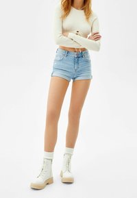 Kvinna som står med armarna i kors, iklädd en beige kortärmad topp med lång ärm, ljusblå denimshorts, vita strumpor och vita snörstövlar.