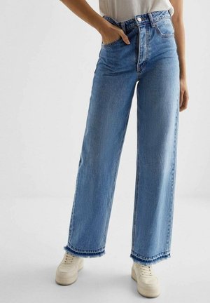 Mannequin portant un jean large bleu clair aux ourlets effilochés, une chemise blanche rentrée et des baskets à plateforme crème. Une main dans la poche.