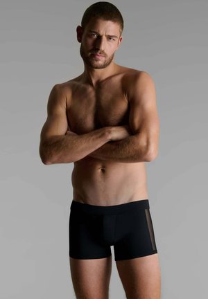 Homme torse nu, aux cheveux courts et à la barbe, les bras croisés, portant un boxer noir devant un fond gris uni.