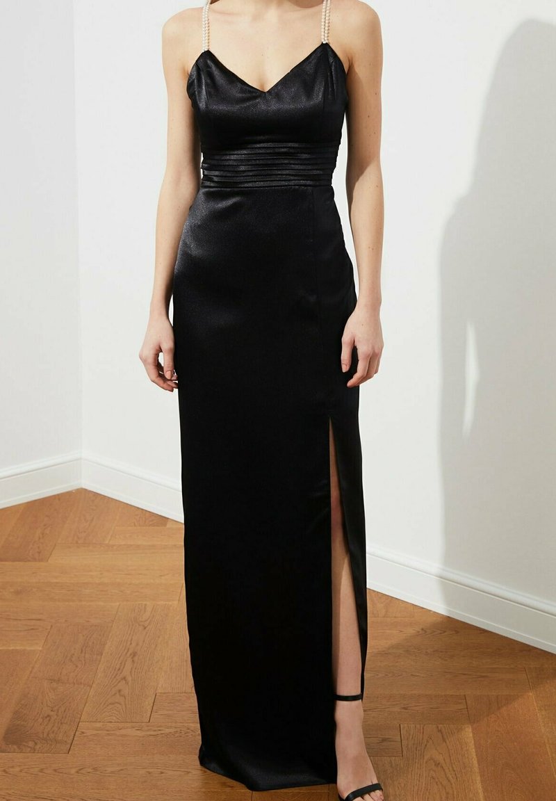 Czarna satynowa sukienka z dopasowaną górą, dekoltem w kształcie litery V i perłowymi ramiączkami. Posiada boczne rozcięcie i gładką fakturę, opadającą do podłogi.
