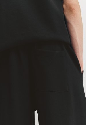 Schwarze Baumwollhose mit lockerem Schnitt, ausgestattet mit einer quadratischen Fronttasche und glatter Textur, die ein minimalistisches Design zeigt.